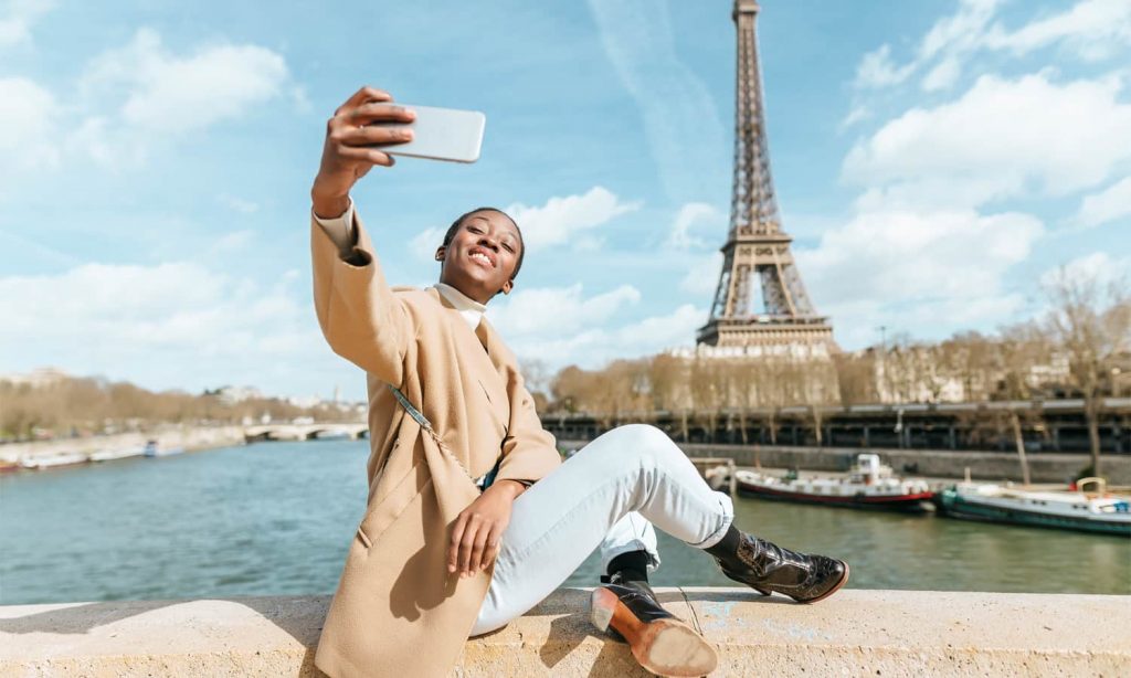 Smiling woman taking a selfie with the Seine River and Eiffel Towel in the background.