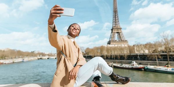 Smiling woman taking a selfie with the Seine River and Eiffel Towel in the background.