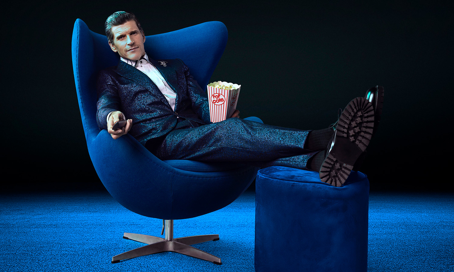 Osher Günsberg in an armchair holding popcorn and a TV remote.
