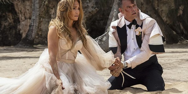 Jennifer Lopez and Josh Duhamel in Shotgun Wedding. They're kneeling on the beach, hands tied together, covered in dirt and blood, after their wedding was ambushed by armed pirates.