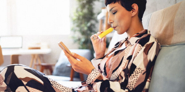 Vape laws in Qld could radically change after the state launched a parliamentary inquiry. Image shows a woman sitting on a sofa using a vape.