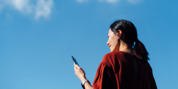 Low angle portrait of young Asian woman using smartphone against cloudy, blue sky.