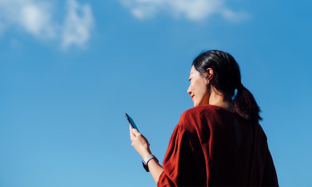 Low angle portrait of young Asian woman using smartphone against cloudy, blue sky.