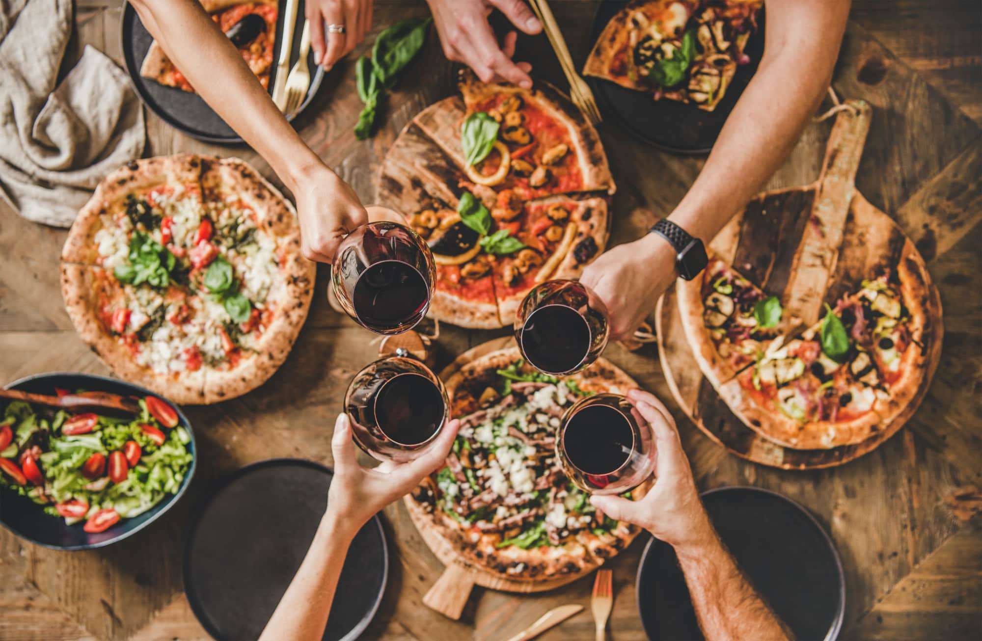 Family or friends having pizza party dinner. Flat-lay of people clinking glasses with red wine over rustic wooden table with various kinds of Italian pizza, top view. Fast food lunch, celebration (Family or friends having pizza party dinner. Flat-lay
